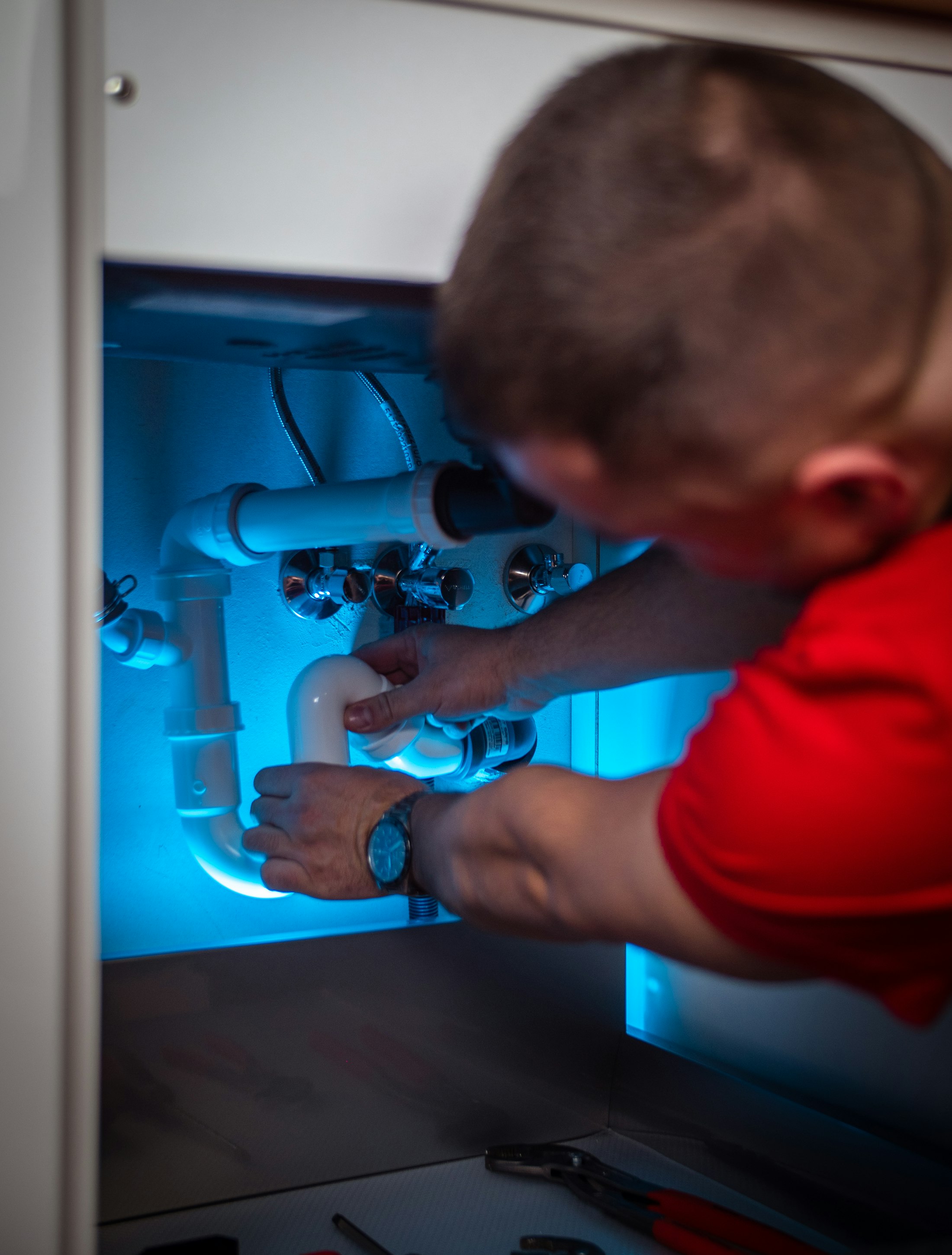 A plumber in a red shirt repairing supply lines under a kitchen sink
